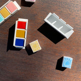 White powder coated metal holders containing brightly coloured watercolour paints, one empty holder and some wrapped watercolour half pans. On a wooden surface.